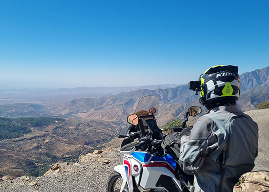 Radu Prie. Overlanding motorcyclist overlooking hills and valleys from high elevation.