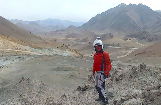 Steve Madden on his RTW motorcycle trip, standing surrounded by rocky trails and mountains.